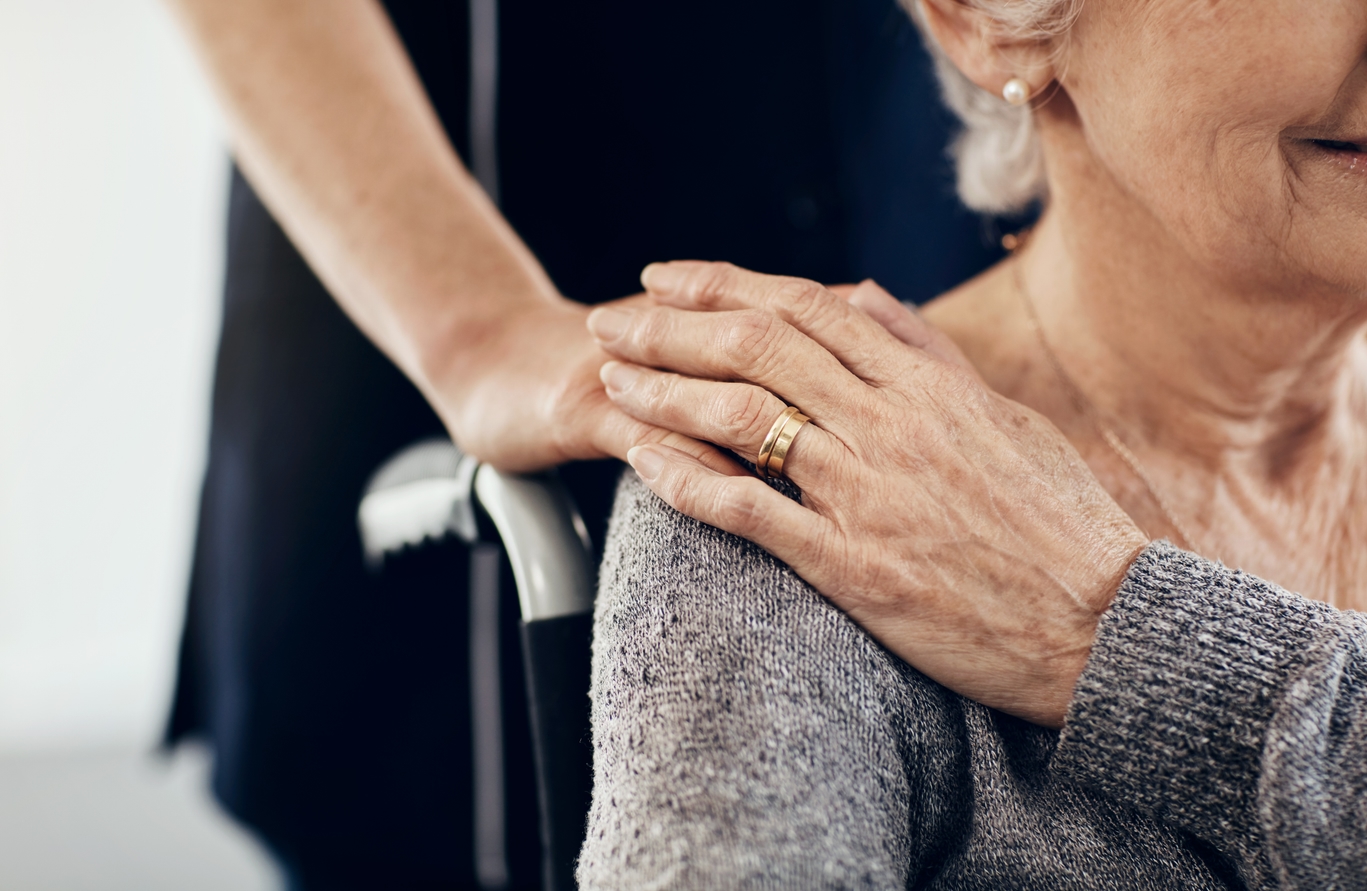 Zorgverlener legt geruststellend een hand op de schouder van een oudere vrouw in een rolstoel